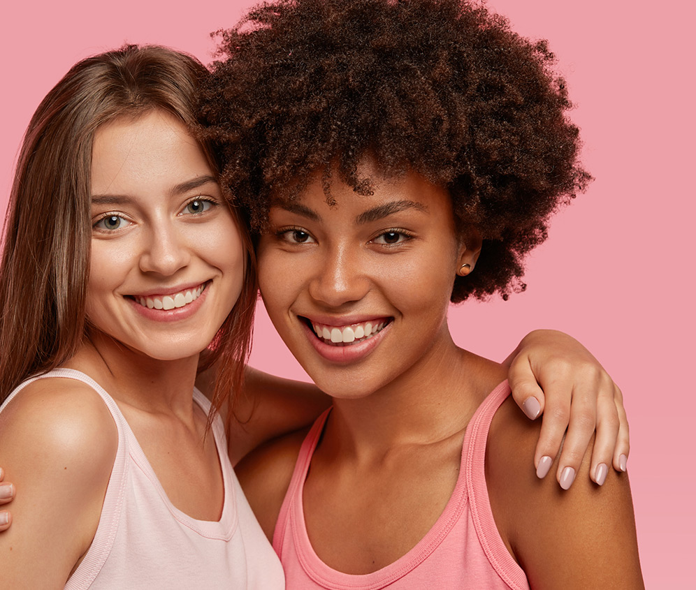 Duas mulheres jovens abraçadas sorrindo para câmera, usando roupas rosas em um fundo rosa mais escuro